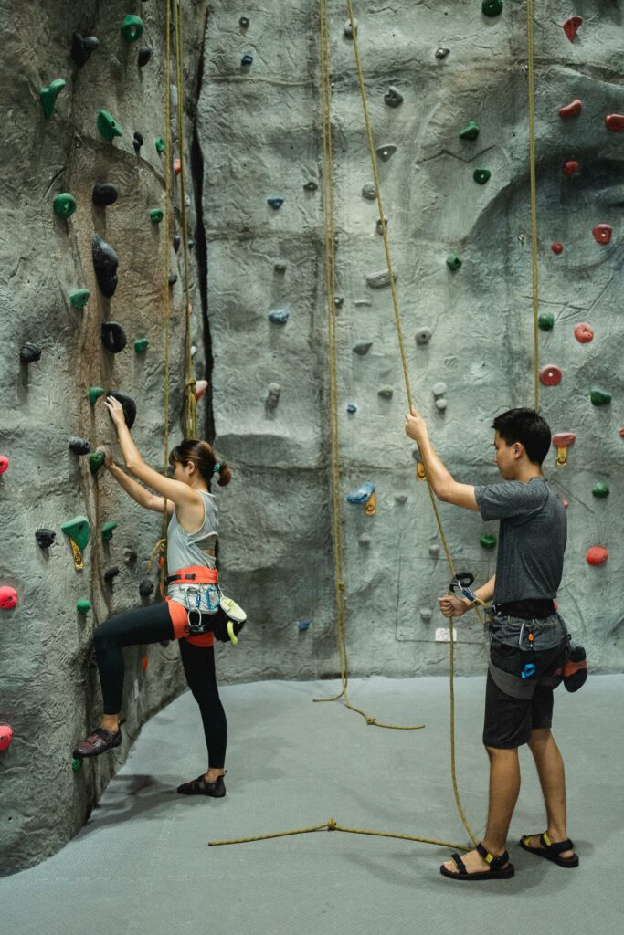 Woman rock climbing indoors with trainer belaying rope for safety.